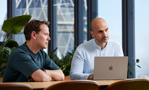 Ahmed hat sein MacBook vor sich und sitzt mit einem Kollegen an einem Tisch. Er schaut jemanden außerhalb des Fotos an.