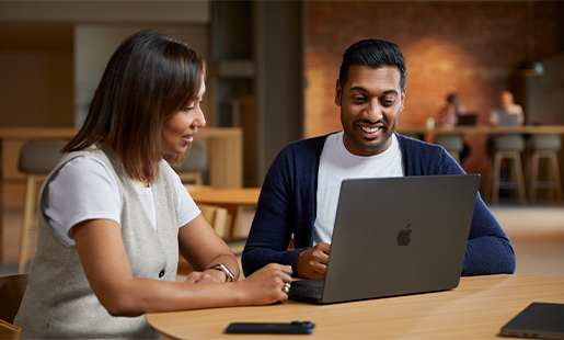 Amar sitzt mit seinem MacBook an einem Tisch in einem Gemeinschaftsbereich und arbeitet mit einer Kollegin zusammen.