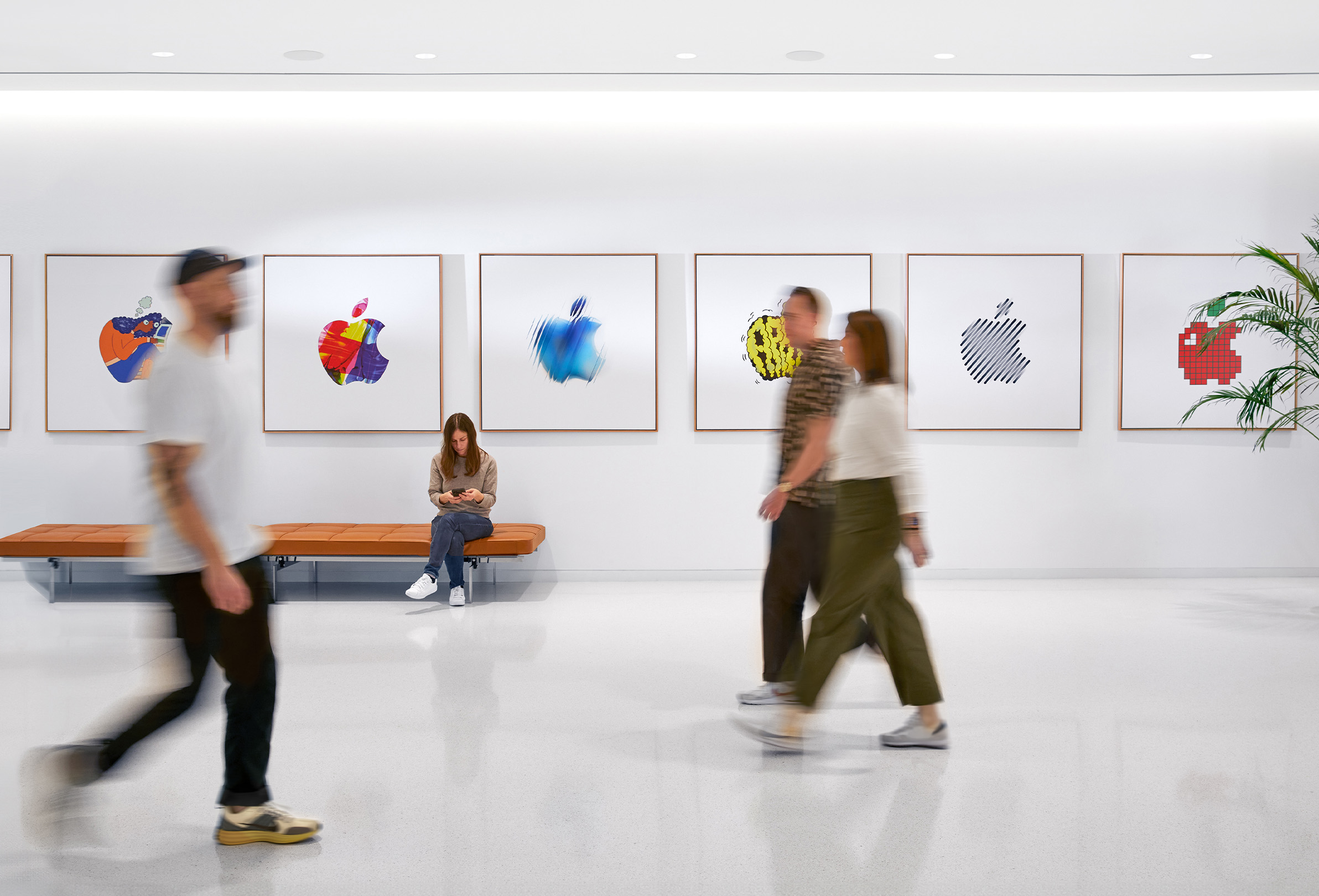 세 명의 Apple 동료들이 컬러풀한 Apple 로고들로 장식된 실내 벽을 지나쳐 가고 있고, 다른 Apple 직원 한 명이 벤치에 앉아 있음.