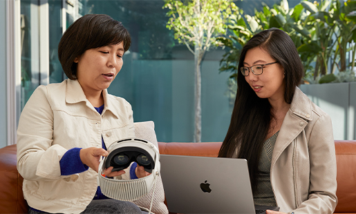 Jessica가 테라스 쪽으로 문이 열려 있는 실내에 앉아서 MacBook을 사용하고 있고, 옆에 앉은 Apple 동료가 Jessica에게 Apple Vision Pro를 보여주고 있음.