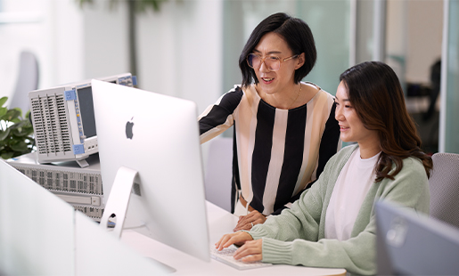 Ella sentada ao lado de um colega que está sentado em frente a um monitor Apple.