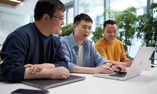 Ken a escrever no MacBook, com um colega sentado em cada lado e plantas e janelas luminosas em segundo plano.