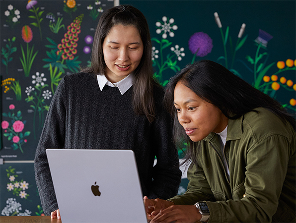 MacBookで一緒に作業している2人のマーケティング関連職のAppleチームメンバー。背後には自分たちのチームがデザインしたポスターが貼られている。