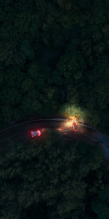 Una vista aérea de una carretera con curvas de noche, dos vehículos ubicados en forma perpendicular con las luces encendidas, rodeados de árboles