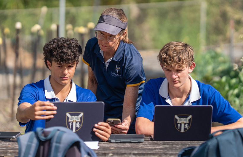 A teacher holds an iPhone and two students review information on their iPad&nbsp;while sitting on a bench in a garden.